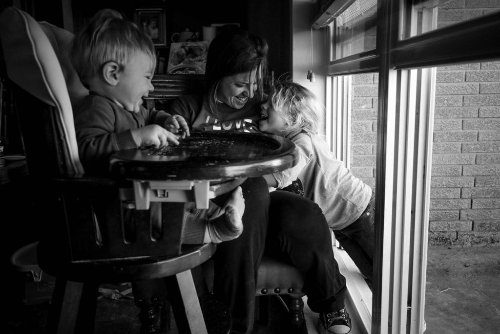 girl leans in through open window to cuddle against mom while baby laughs in high chair