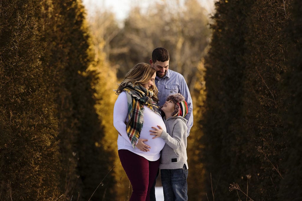boy in tuque looks up at mother while touching her pregnant belly as father looks on while standing near cedars during cornwall family photography session