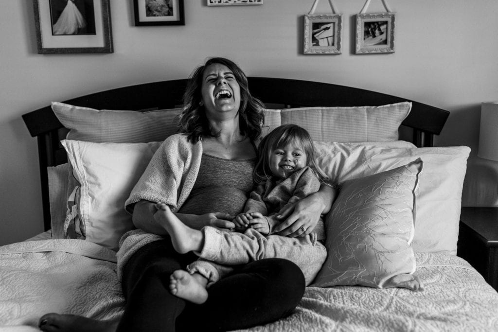 pregnant woman sits on bed and laughs while holding daughter during cornwall family photography session