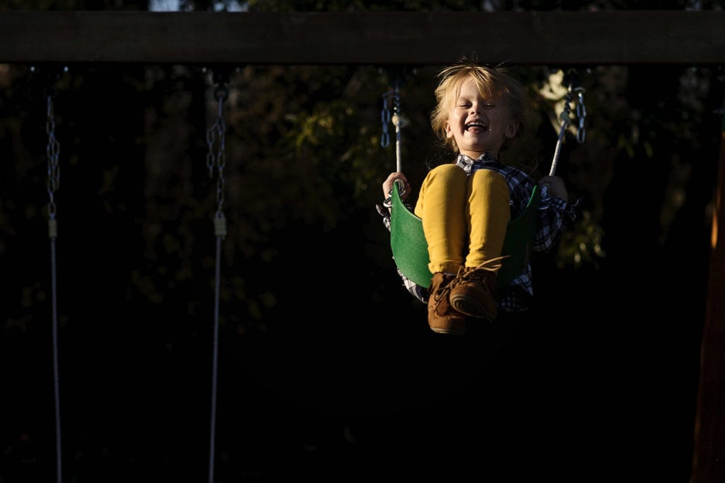 little girl in yellow pants smiles while swinging high on green swing
