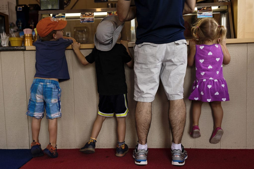 three children line up with adult at ice cream counter while little girl pulls herself up to see inside freezer