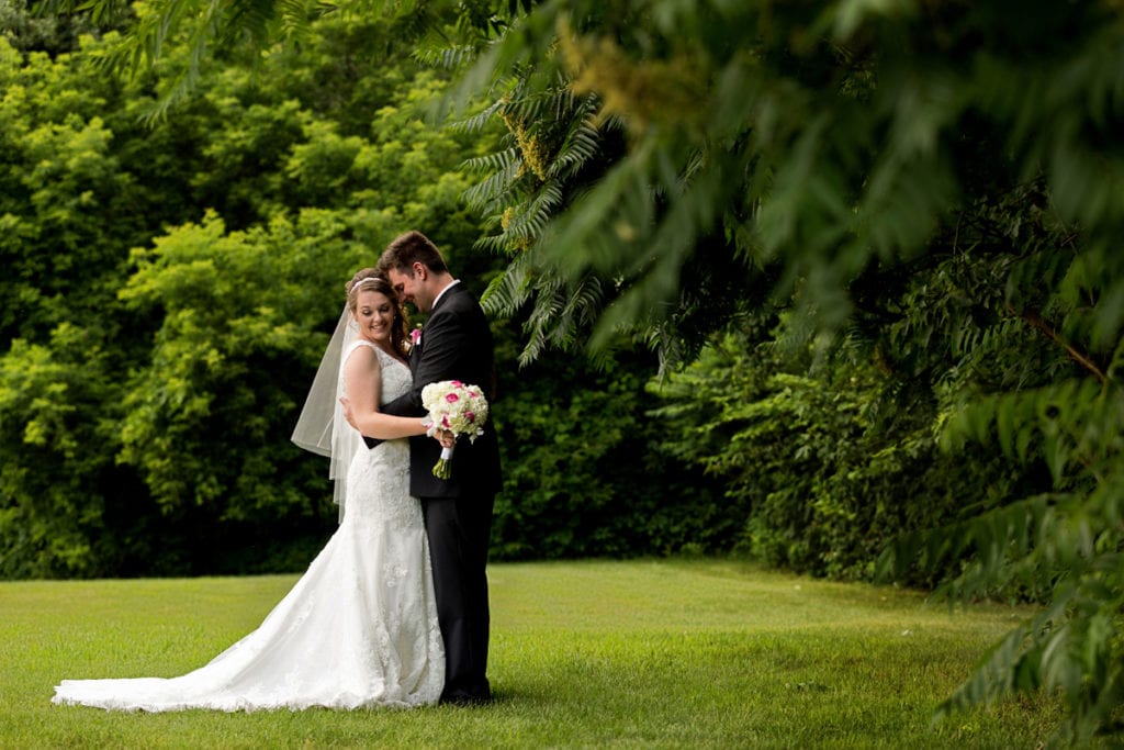 Bride holding bouquet and groom with arms around her waist in lush greenspace