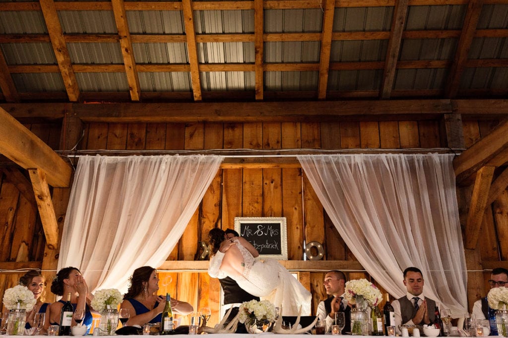 Groom picking up bride at head table with wedding party cheering