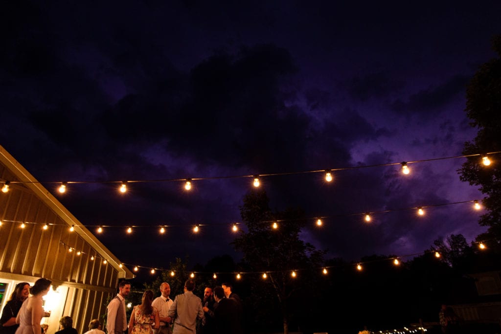Wedding reception guests on outdoor patio under string lights and dramatic clouds with lightning