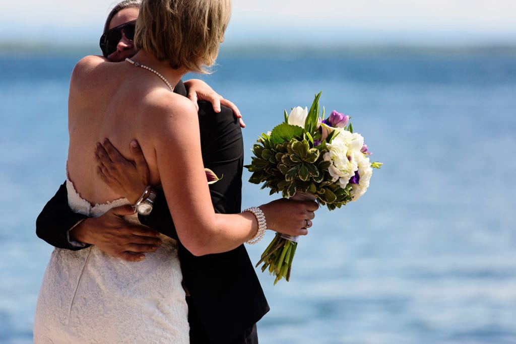 bride in pearl necklace hugging bride in suit on riverfront dock