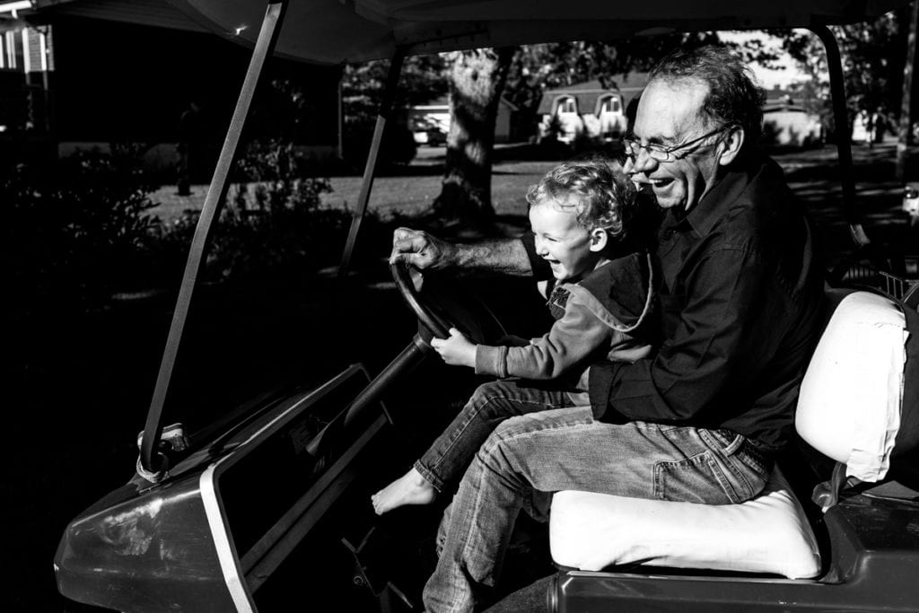 grandpa and little boy laughing while driving golf cart during candid Cornwall backyard family session