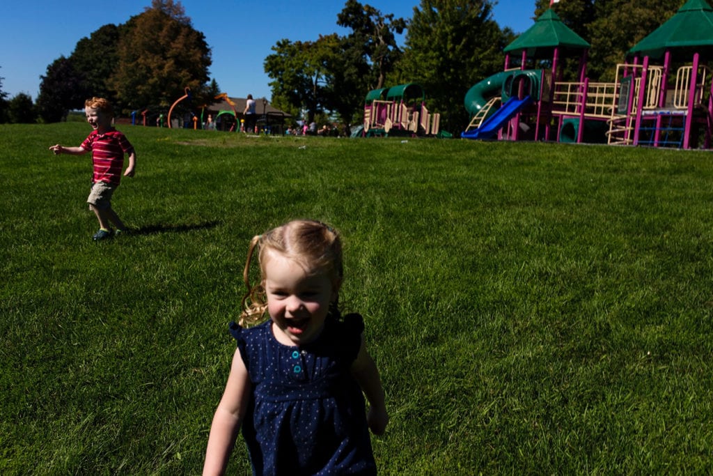 little boy and girl laughing and smiling while running down grassy hill in front of park