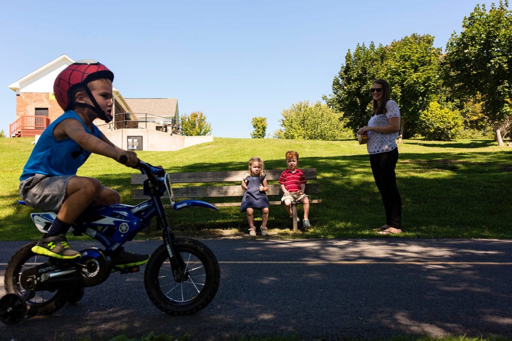 brother and sister sitting on park bench while young boy bikes past during candid Cornwall outdoor family session