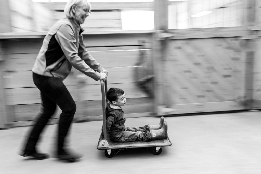 grandma playing and pushing grandson on wheeled cart in horse stable