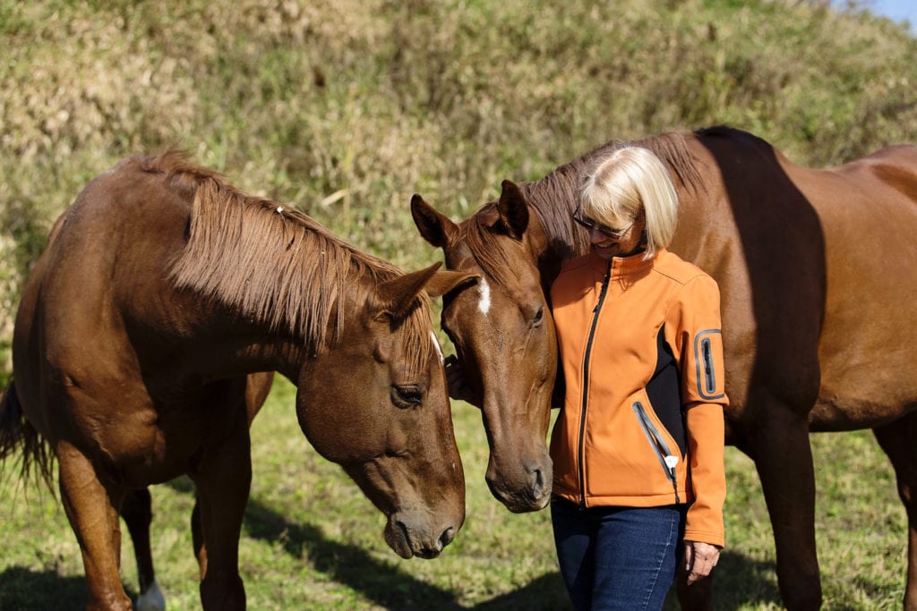 woman in bright orange zip up sweater with two horses in storytelling photo session