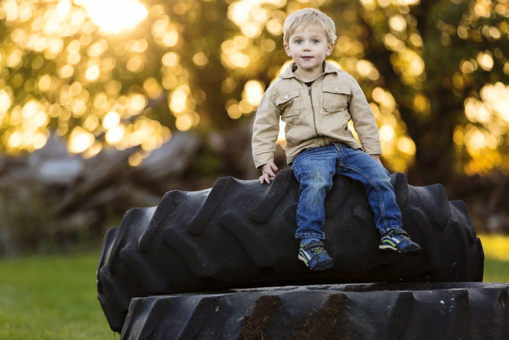 young boy in jeans and jacket sitting on tractor tire in golden hour fall family session