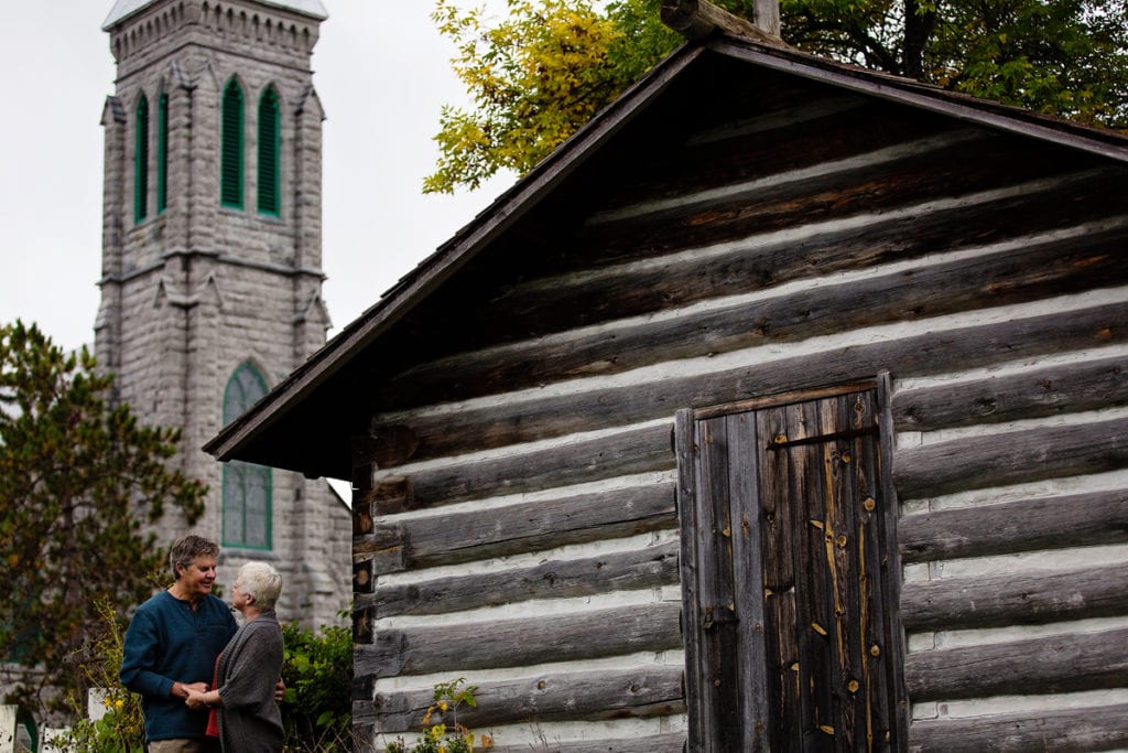 Couple beside old log cabin with church in background during Ontario Casual Family Portraits