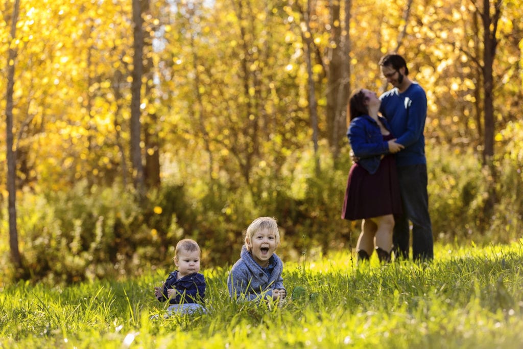brother and sister sitting in grass laughing with parents in sunlit nature background