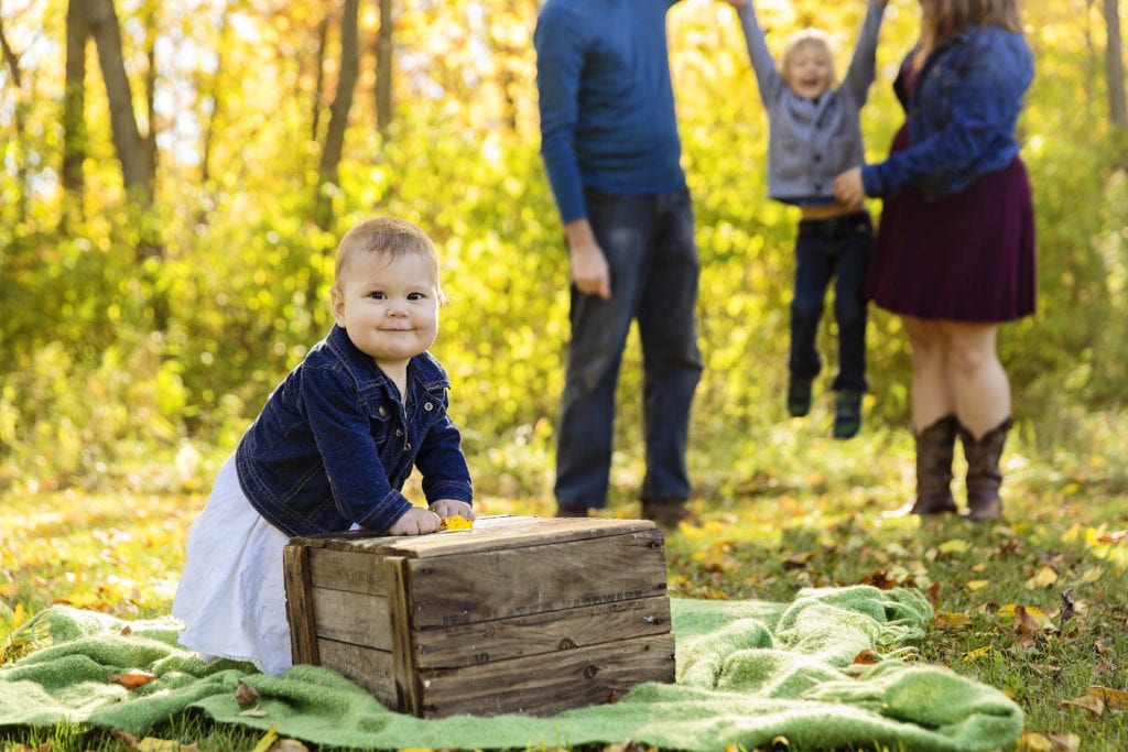 baby girl leaning on wooden box with family playing in treed background