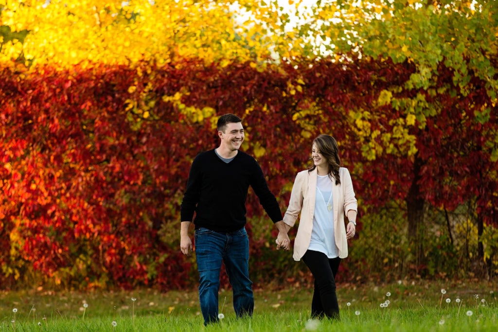 couple holding hands and laughing in front of fall foliage during Autumnal Cornwall engagement session