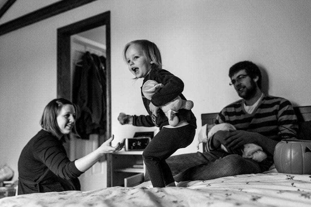 daughter jumping on bed while father holds newborn son