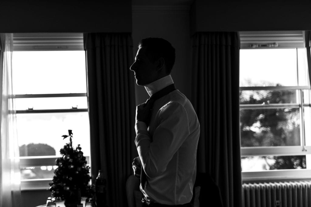 groom ties tie in dramatic window light during groom prep at Christchurch Habour Hotel & Spa