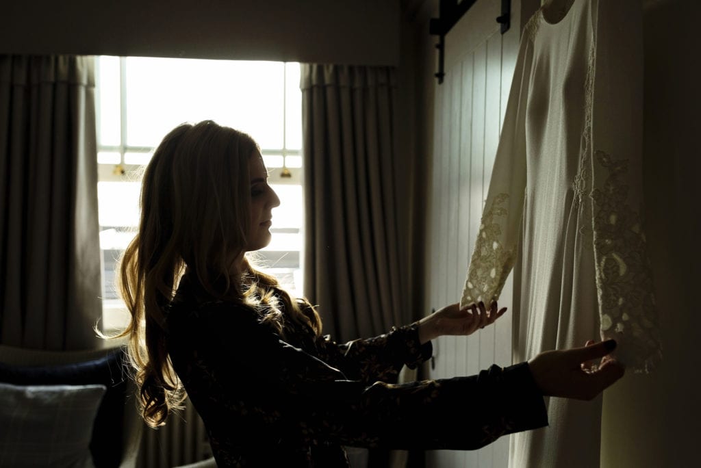 bride holds out sleeves of dress and looks at bespoke wedding gown in backlight from window during bridal prep at Christchurch Habour Hotel & Spa
