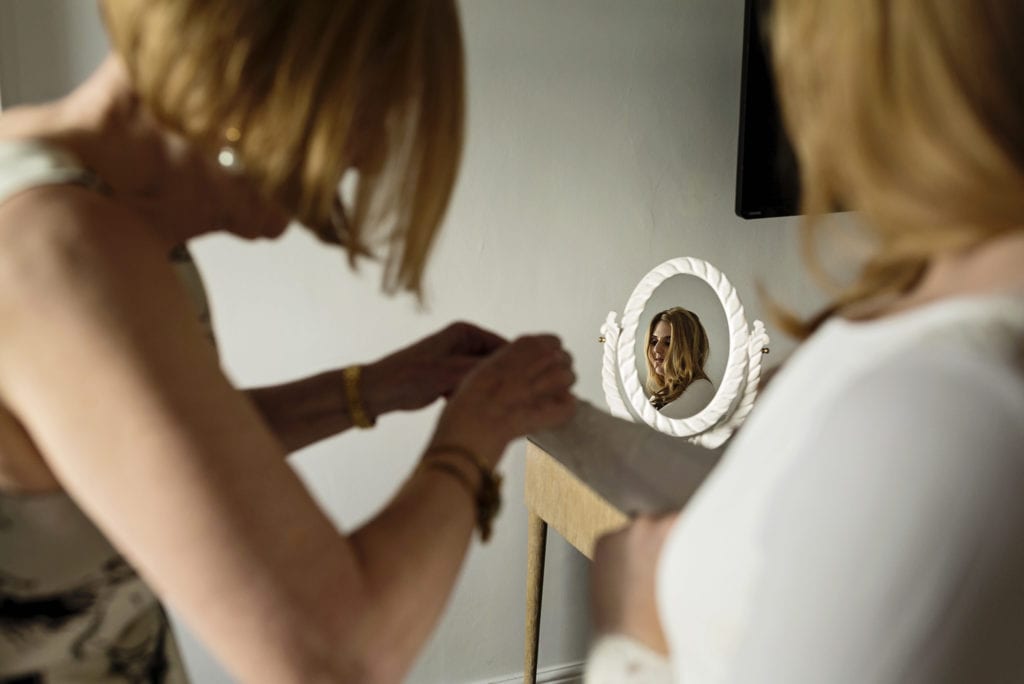 mother of the bride fastens sleeve buttons on bespoke bridal gown while bride's face shows in mirror during bridal prep at Christchurch Habour Hotel & Spa