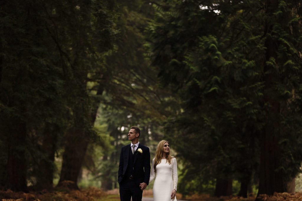 bride and groom hold hands while standing on road and looking up at trees on Rhinefield Ornamental Drive during chic New Forest wedding portraits