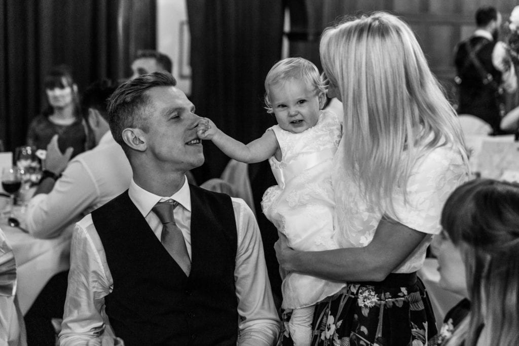 woman stands and holds little girl in white dress who squeezes groom's nose during wedding reception at Rhinefield House