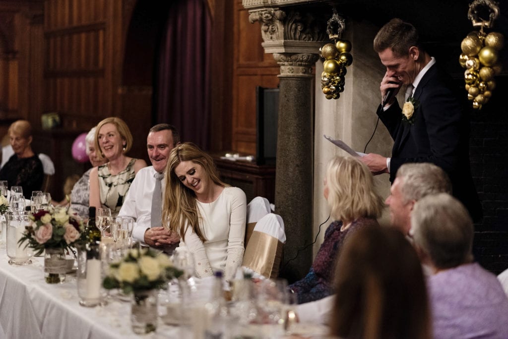 Bride looks down bashfully while groom gives speech during wedding reception at Rhinefield House