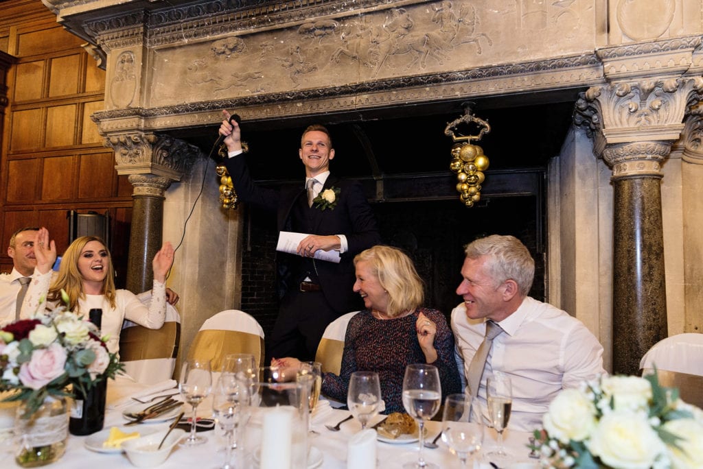 Groom holds mic in the air during speech while bride waves arms in the air from her seat during wedding reception at Rhinefield House