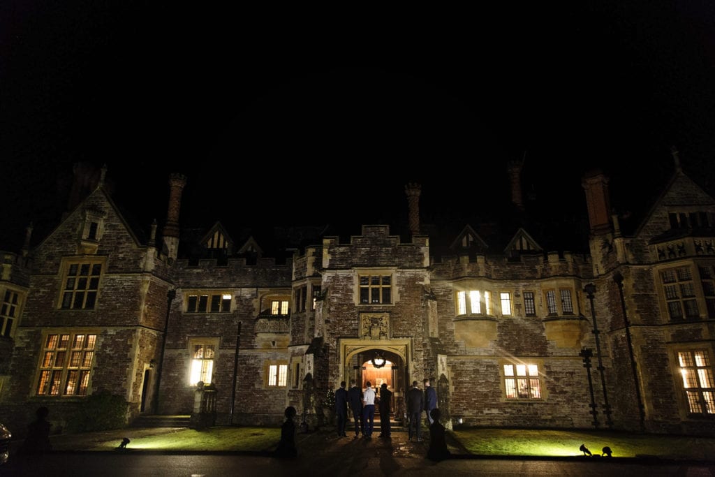 Men stand in front of entrance late into evening during wedding reception at Rhinefield House