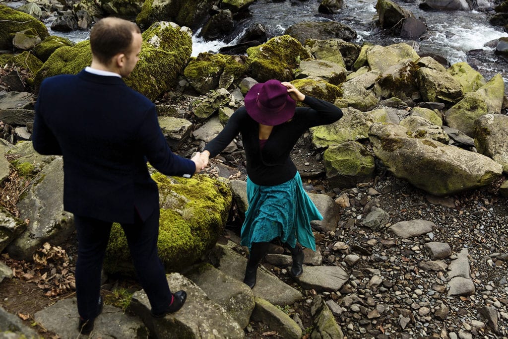 Man gives wife in purple hat a hand while walking up stone steps by river