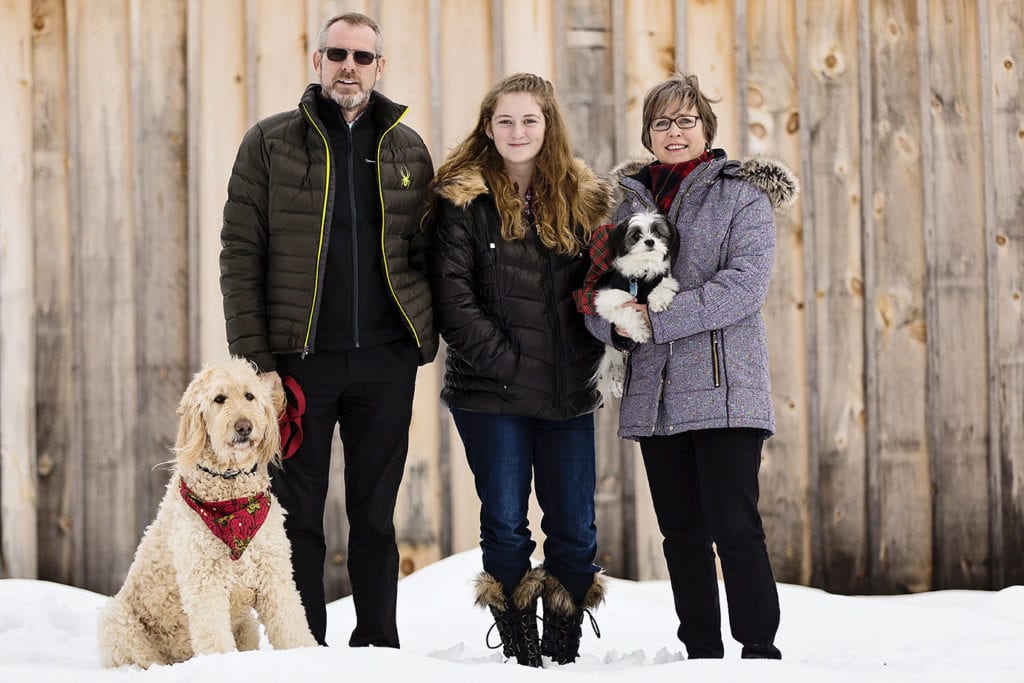 family with large and small dog stand in front of wood shed