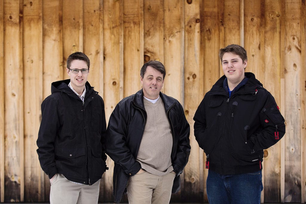 man stands in front of raw wood wall with two sons all in black jackets