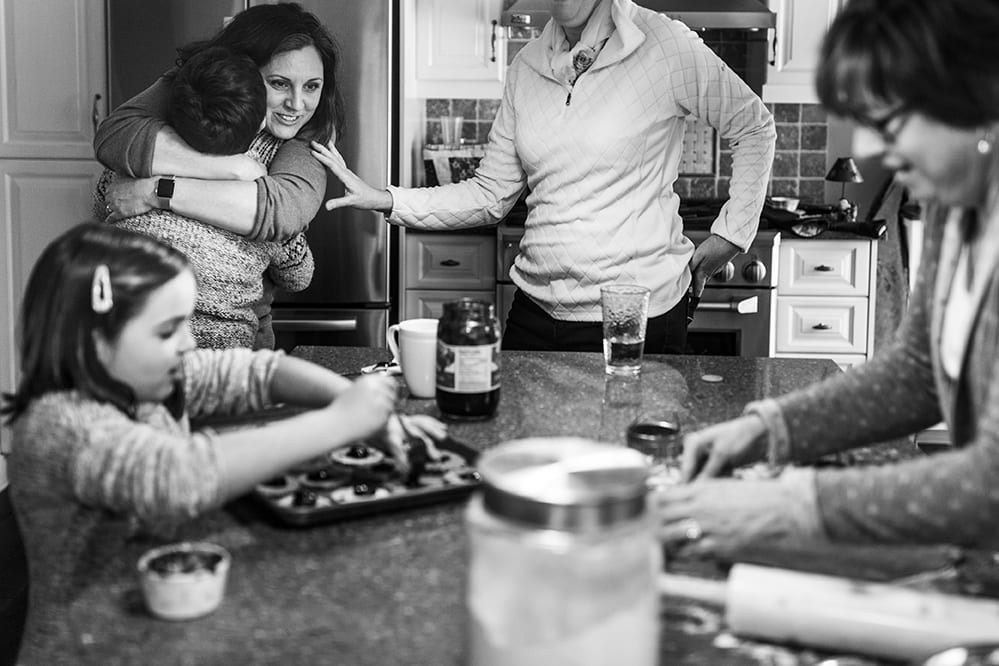 mother hugs son while sister touches her arm and daughter bakes with grandmother
