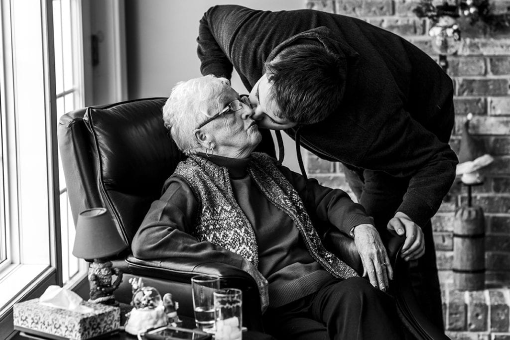 Grandson bends over grandmother's chair to kiss her on the cheek