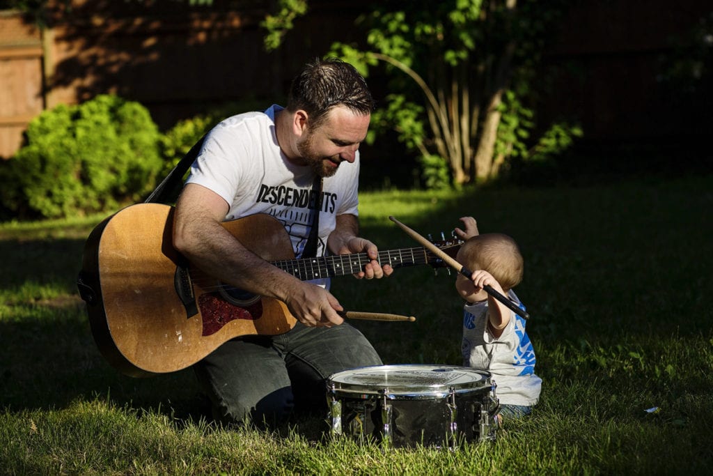 father with guitar helps son play drums in sunshine