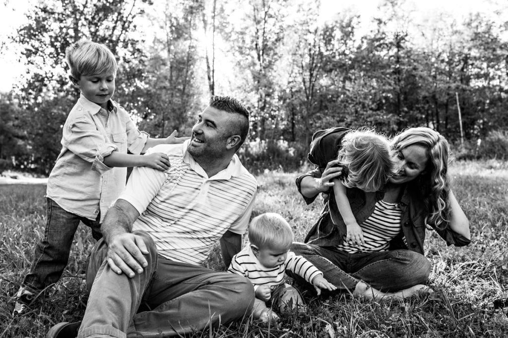 three boys climb on parents who are sitting in grass during session with Ontario family photographer