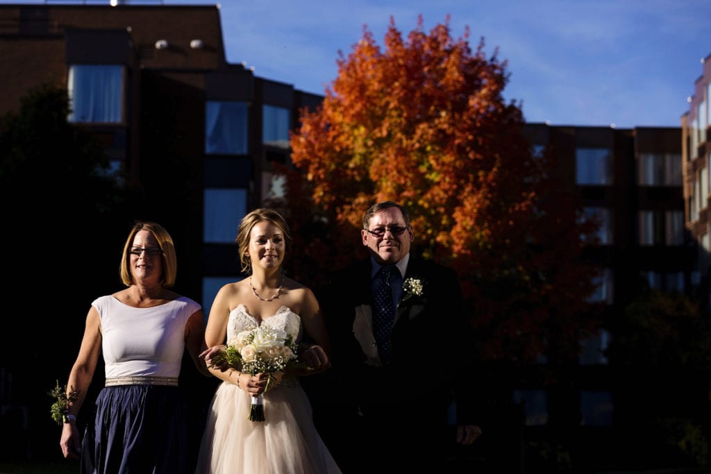 bride makes ceremony entry with parents in dramatic sunlight for Autumnal NAV Centre Cornwall Wedding