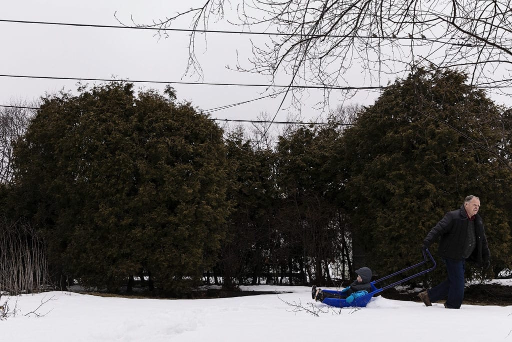 boy rides around yard on shovel while being pulled by grandpa during during Ontario Family Photojournalism session