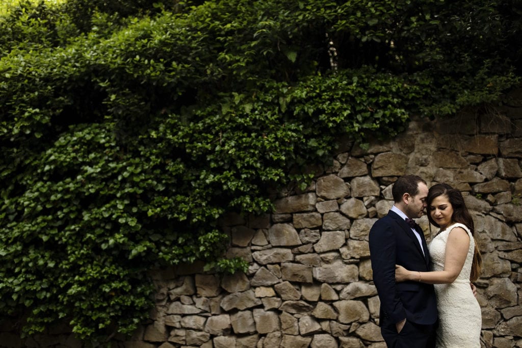 bride and groom snuggle in front of stone wall covered with ivy during session with Ontario & Destination Wedding Photographer