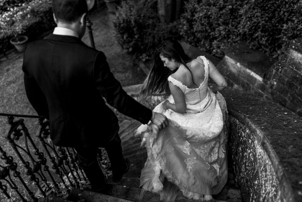 bride walks down sweeping staircase while groom picks up back of fit and flare gown during session with Ontario & Destination Wedding Photographer
