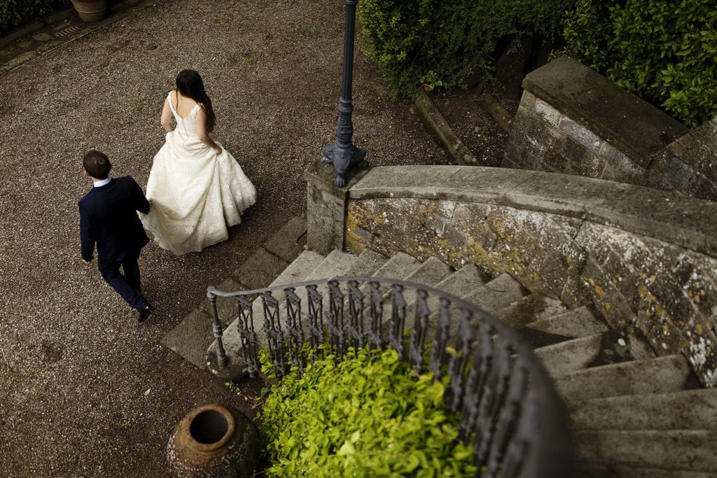 bride walks away from sweeping Italian staircase while groom carries back of her gown during session with Ontario & Destination Wedding Photographer