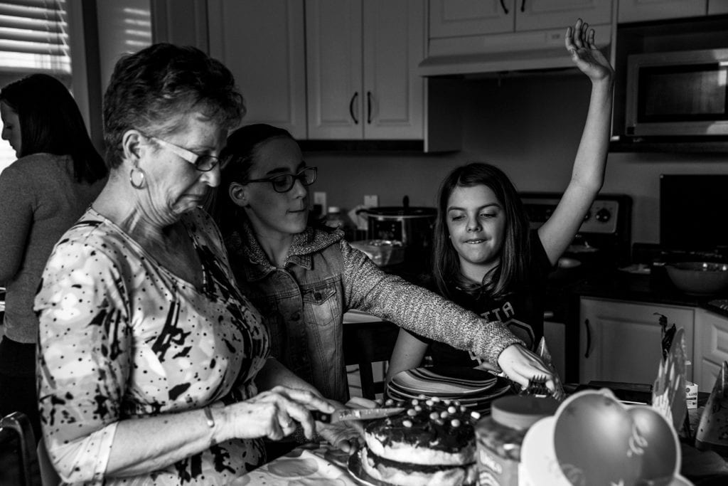 grandma slices cake while granddaughter raises hand to show she wants some