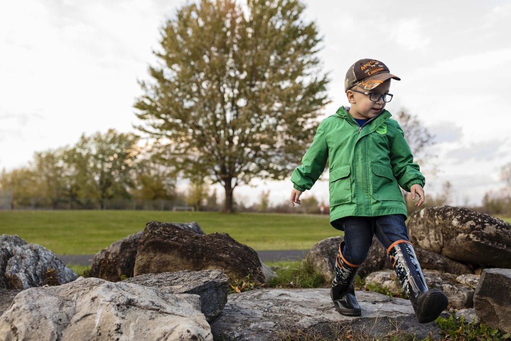 boy in rubber boots walkes over rocks with fall colours in tree behind him