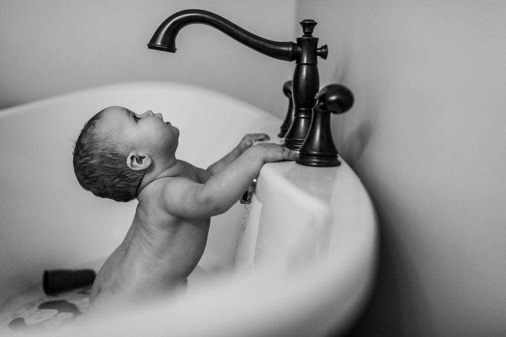 baby stands in tub and looks up at tap