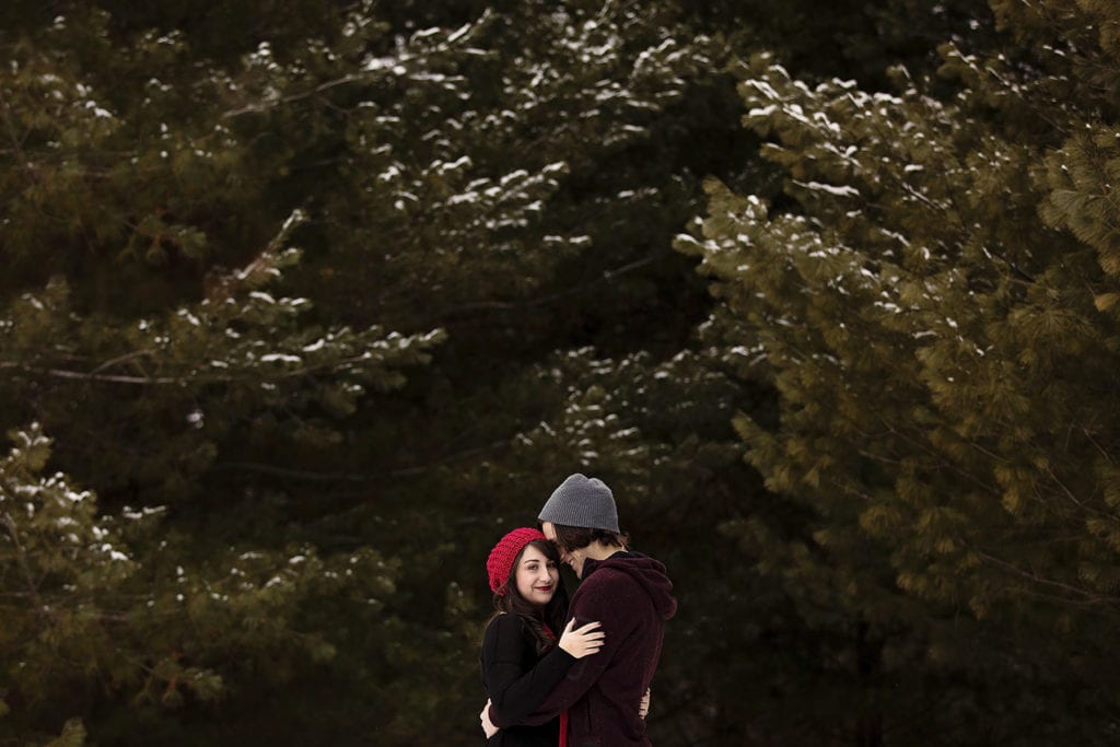 smiling couple embracing in snow kissed pine grove during winter engagement session