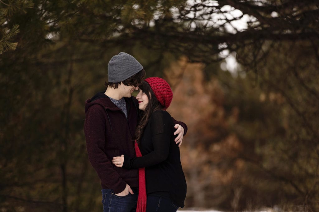 romantic couple cuddling in pine trees during winter engagement shoot