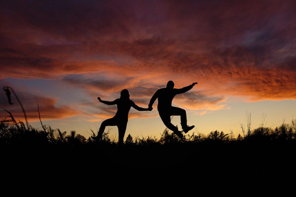silhouette of couple jumping for joy against sunset sky in Cornwall Autumn Engagement Session