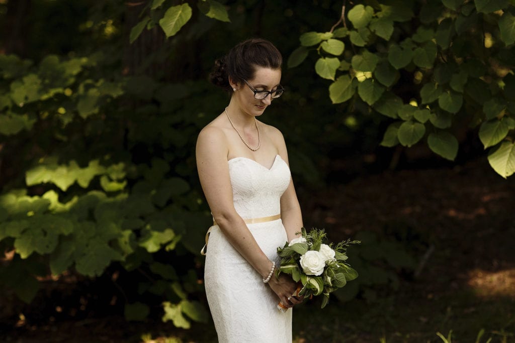 classic bride wearing glasses holding white rose bouquet in natural backdrop at intimate ottawa park wedding