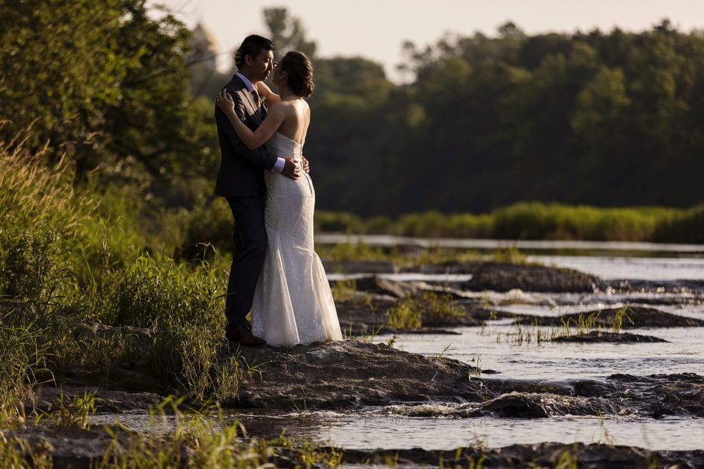 bride and groom embracing on shore of river during intimate ottawa park wedding