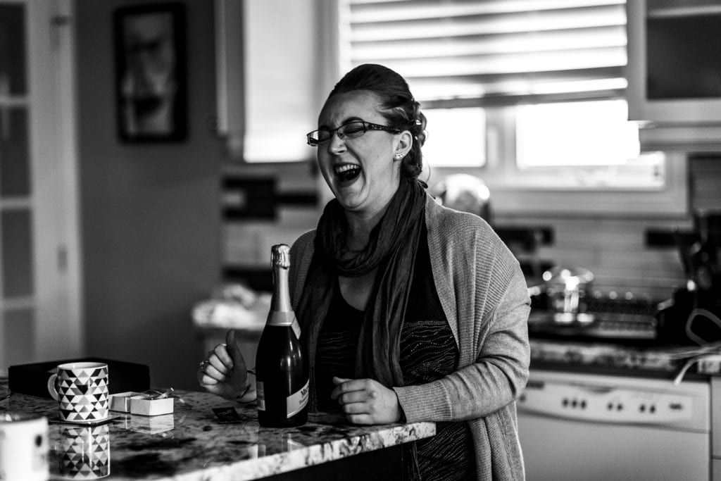 woman laughs enthusiastically while standing at kitchen counter in front of bottle of sparkling wine