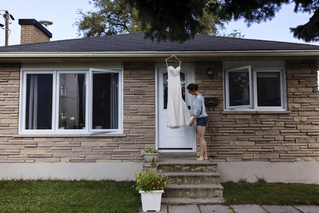 bride adjusts wedding dress while it hangs from eavestrough